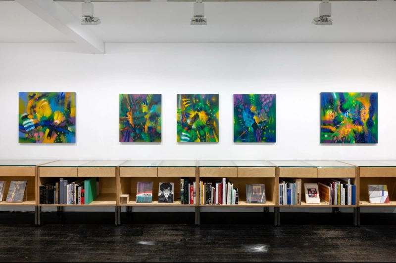 Five colorful abstract paintings displayed above a long wooden bookshelf in a gallery.