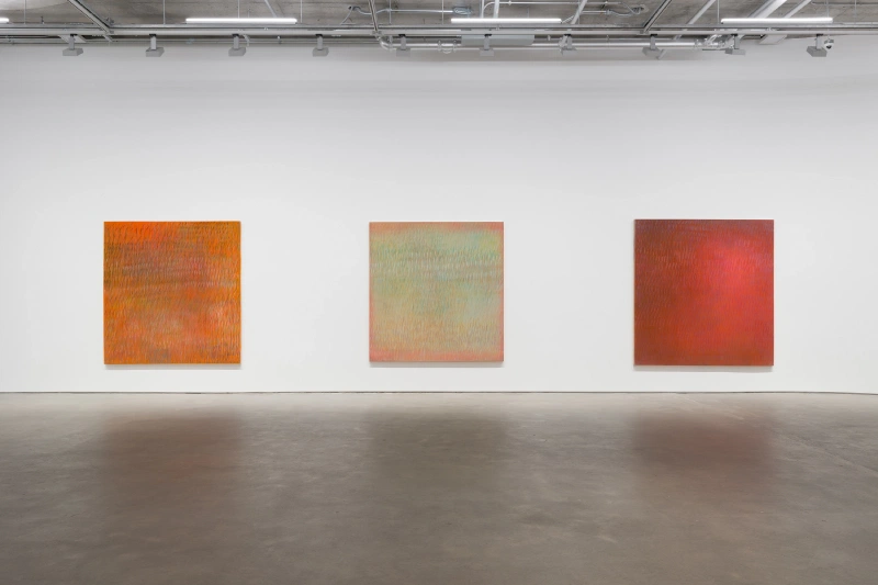 Three square color-field paintings in orange, pale green, and red displayed on a white gallery wall.