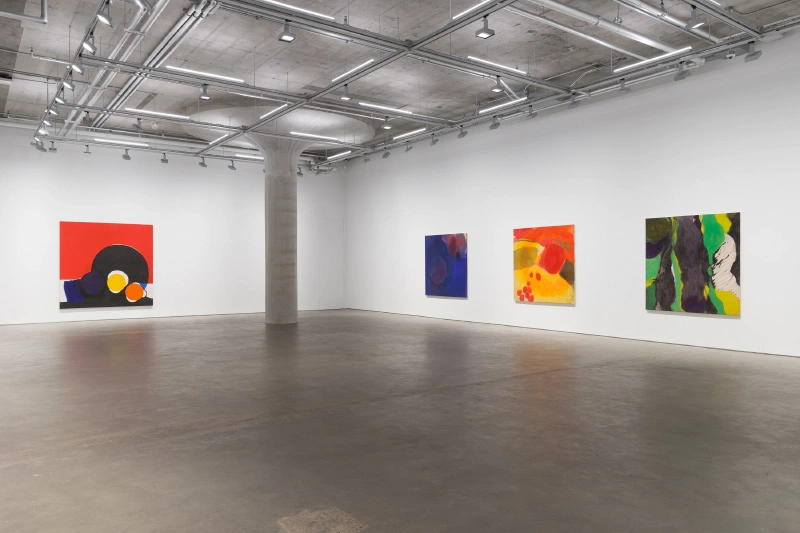 Four vivid geometric abstract paintings displayed around a concrete column in a gallery.